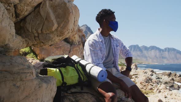 African american man wearing face mask hiking in mountain countryside sitting on a rock