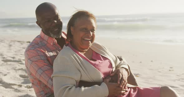 Smiling senior african american couple embracing and sitting on sunny beach