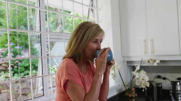 Caucasian senior woman standing in kitchen at home drinking cup of coffee