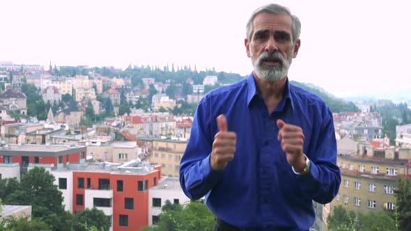 Old Senior Man Talks To Camera - City (Buildings) in Background