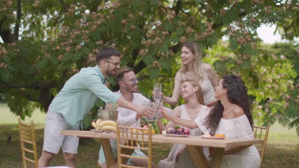 Group of happy young people cheering with drinks and eating fruits by the pool in the garden