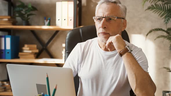 Senior Businessman is Looking Thoughtful Sitting at Desk and Typing on His Laptop While Working at