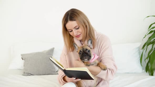 Young Beautiful Woman in a Cozy Powder Pink Sweater and White Stockings Sits on the Bed with Her Dog