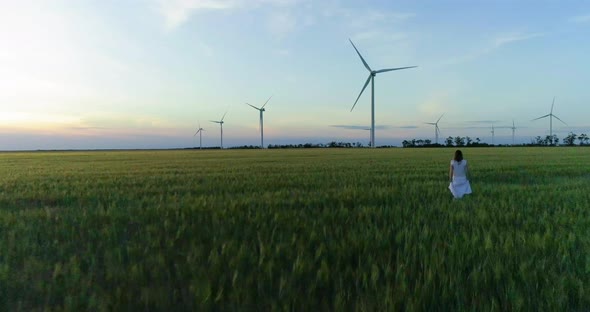 Beautiful girl walking on a green wheat field with windmills for electric power production