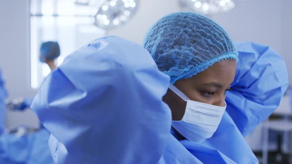 African american female surgeon wearing surgical cap, putting face mask on in operating theatre