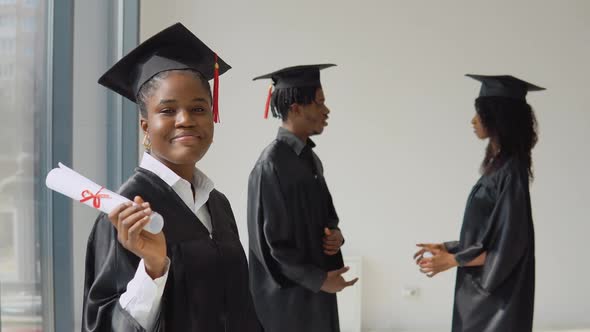 One Young AfricanAmerican Woman Stands in the Foreground and Looks at the Camera with a Diploma in