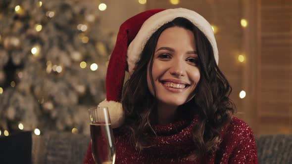 Attractive Woman with a Glass of Champagne in Santa Hat Looking at the Camera on Christmas Tree