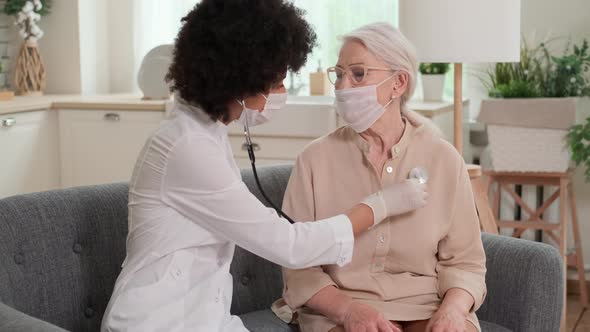 Afro American Woman Doctor in Mask Listens to the Breathing of an Elderly Woman at Home and Supports