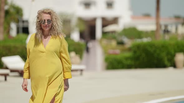 a Curly Blonde in a Yellow Dress Goes and Adjusts Sunglasses Against the Background of Blurred Sun