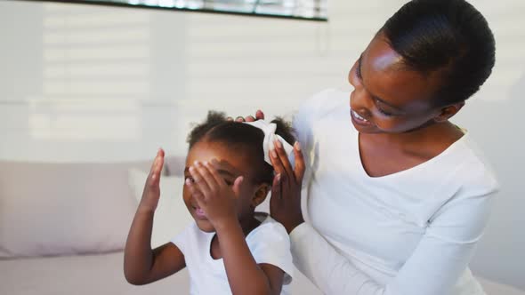 Happy african american mother and daughter sitting on bed, mother putting hair bow