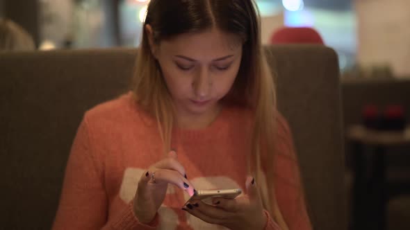 Young Beautiful Girl Sits in a Cafe Works on a Smartphone