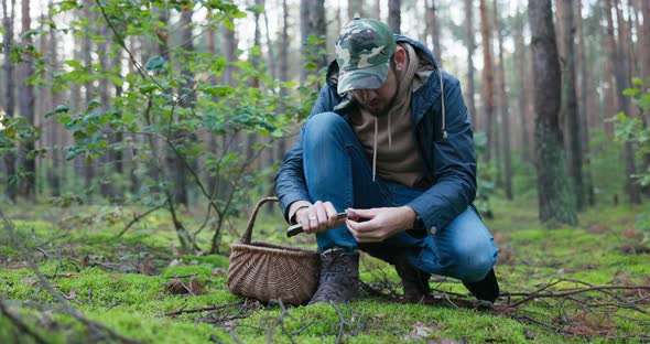 Mushroom Picker Dressed in Jacket and Helmet Walking in Forest Leaning Close to Ground Puts Basket