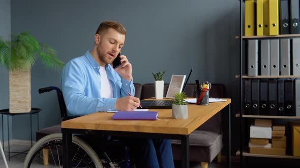 Motivated Handicapped Freelancer Talking on the Phone and Taking Notes Sitting in Wheelchair at