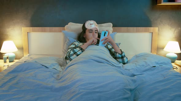 Girl in a Sleep Mask on Her Head at Night Lying in Bed and Using Her Cell Phone