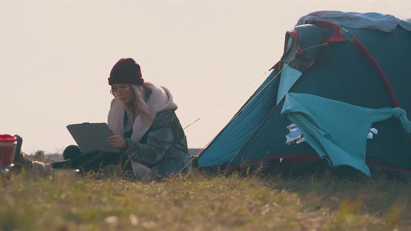 Girl Tourist in Red Hat Sits with Tablet By Tent at Sunset