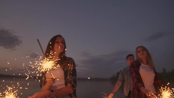 Cheerful Male and Female Friends are Running Along the Beach at Sunset Holding Sparkling Fireworks