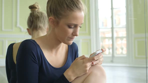 Female Ballet Dancer crouches in front of the studio mirror and uses her smartphone