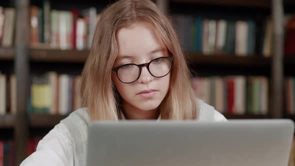 Studying Online Teen Girl Wearing Glasses Looking Laptop Typing Message Over Library Background