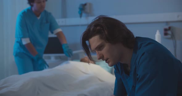 Stressed Young Man Nurse Sitting in Hospital Ward with Colleague Covering Dead Patient with Sheet