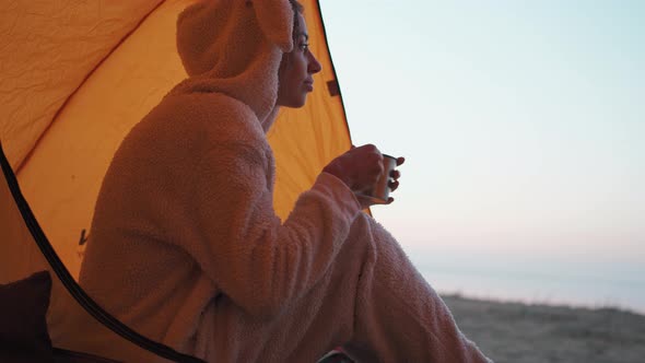Pretty Woman in Funny Pajama Like Bunny Sitting at Tent Entry and Drinking Hot Coffee From Metal Mug