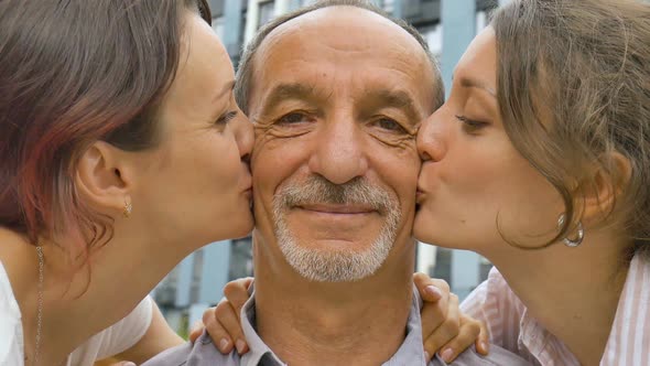 Family Portrait of Senior Father and Two Adult Daughters Kissing Him Outdoors on Modern Building of