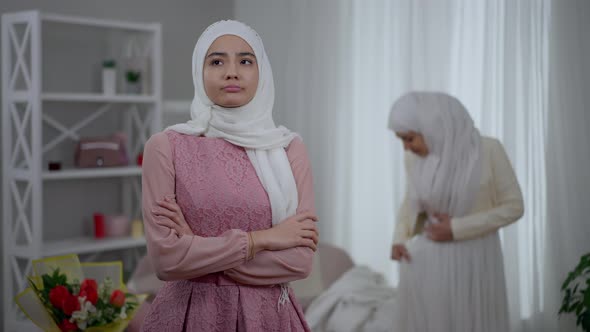 Young Jealous Middle Eastern Bridesmaid with Crossed Hands Looking at Happy Bride Trying on Wedding