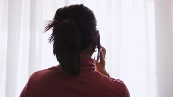 Asian woman talking on cell phone near window with sunlight.