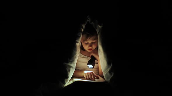 Little Girl is Read a Book Under a Blanket with a Flashlight