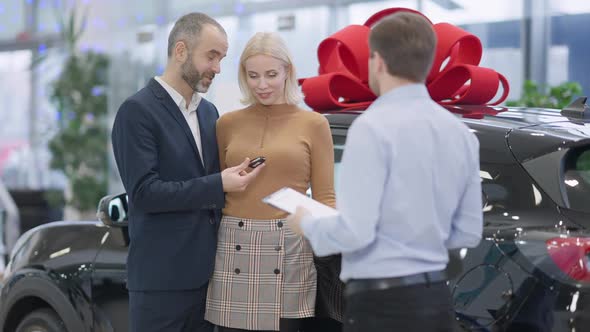 Loving Caucasian Man Giving Car Key To Smiling Beautiful Woman in Showroom with Blurred Dealer