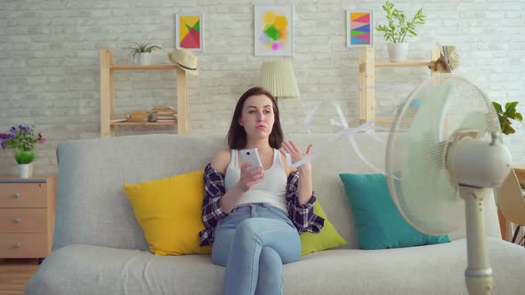 Young Woman Uses a Smartphone Sitting on the Couch in Front of an Electric Fan Escapes From the Heat