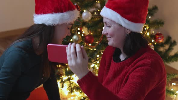 Two Females in Green Santa Hat Using Smartphone on Christmas Tree Background