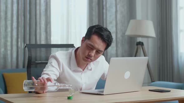 Asian Man Pouring Vodka In A Shot Glass Before Drinking And Typing On A Laptop At home