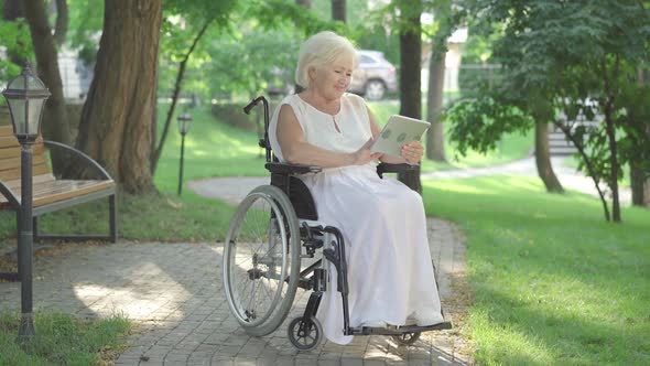 Wide Shot Portrait of Cheerful Disabled Caucasian Woman in Wheelchair Waving at Tablet Selfie Camera