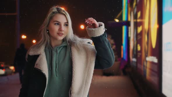 Girl Walks and Smiles Winter Night During Snowfall Near Glowing Shopping Center