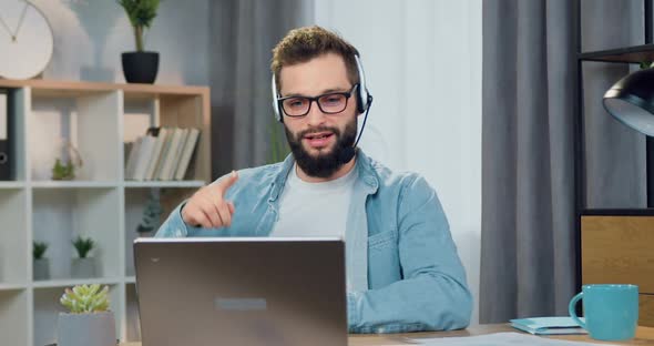 Bearded Worker in Headset Sitting in front of Laptop During Video Meeting