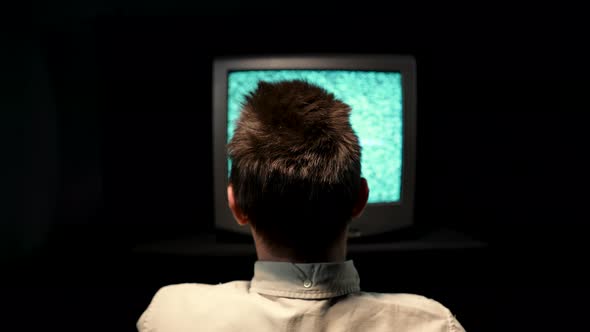 Rear View of a Man Sitting in Front of an Old TV with Noise Interference on a TV Monitor in a Dark