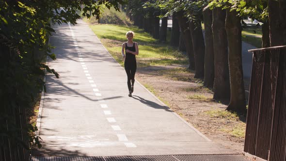 Slim Middle Aged Blonde Woman in Black Sports Bra and Leggings Sportwear Jogging Runs in Summer City