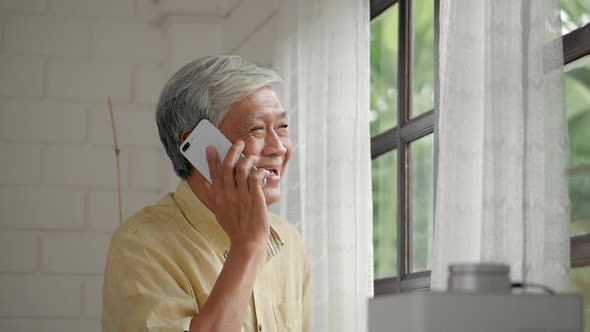 Portrait senior Asian man talking on smartphone happy and smiling beside a window at home.