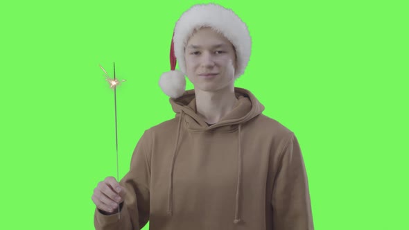 Portrait of Caucasian Teenager in Christmas Hat Holding Sparkler. Happy Boy Posing at Green