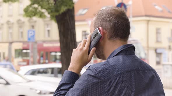 A Caucasian Man Talks on a Smartphone in an Urban Area  Rear Closeup  a Colorful Street