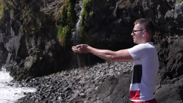 A man on the beach pouring the black sand from his hands under a waterfall