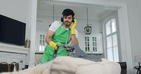 Worker of Cleaning Company Vacuuming Dust Mites on Soft Couch and Enjoying Modern music