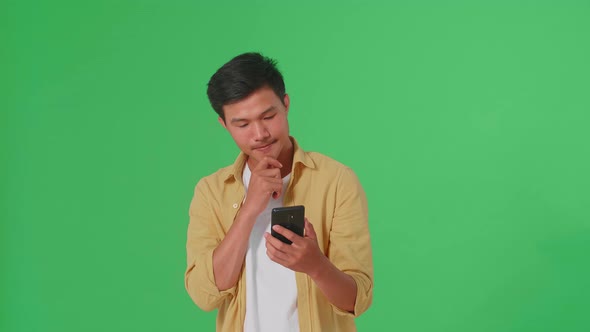Young Asian Man Use Mobile Phone And Thinking About Something In Green Screen Studio