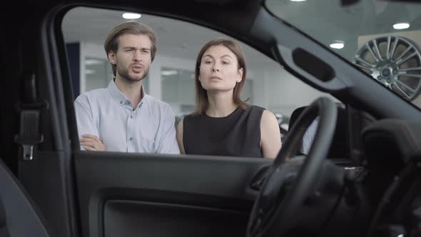 Young Confident Couple Examining New Vehicle in Car Dealership or Showroom and Talking. Portrait of