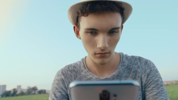 Cute boy in a hat reading messages on his cell phone, blue sky background, camera tracking