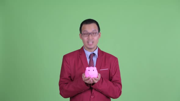 Face of Happy Asian Businessman with Eyeglasses Holding Piggy Bank and Looking Surprised