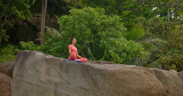 Woman Meditating on the Rock Against Tropical Forest  She Wears Pink Sportswear and Use Yoga Mat