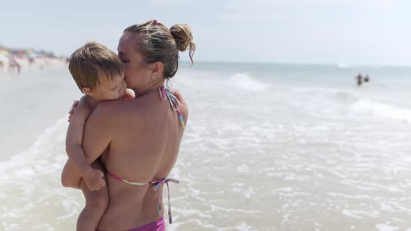 Mom in a Summer Bikini Holds a Baby Who Wants to Sleep on the Beach Near the Sea on Vacation