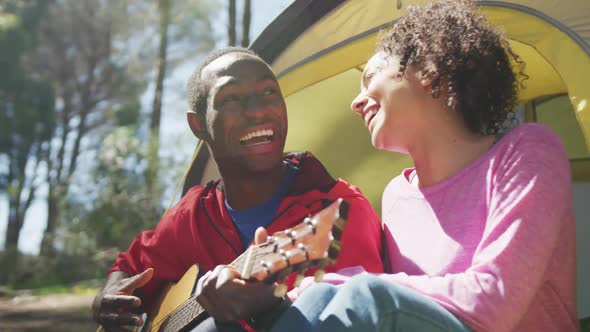 Smiling diverse couple sitting in tent and playing guitar in countryside