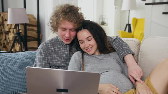 Pregnant Couple Relaxing on the Comfortable Sofa at Home and Watching Family Photoes on Laptop